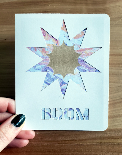 A folded card with a 10-pointed star cut out of it to reveal a different color of paper which in turn has been engraved with another 10-pointed star.