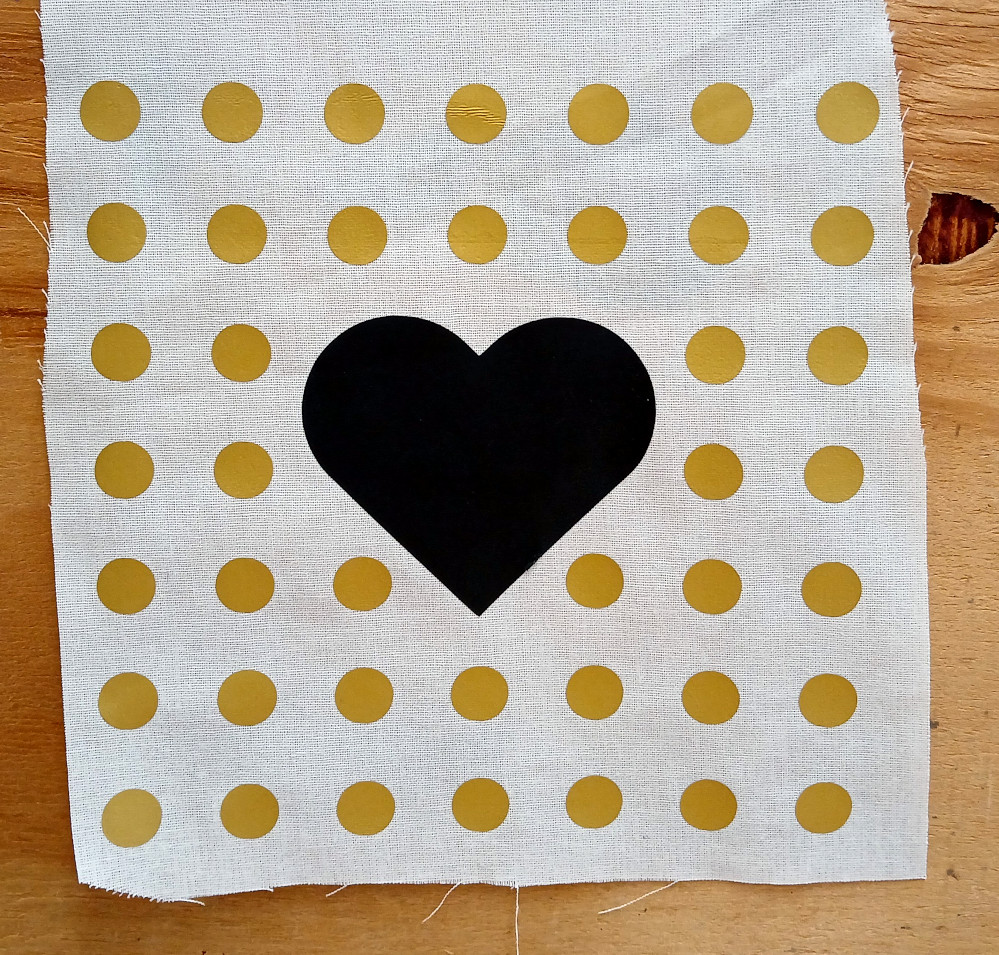 A grid of gold vinyl dots with a precisely aligned black vinyl heart in the center, all of which are on a fabric background.
