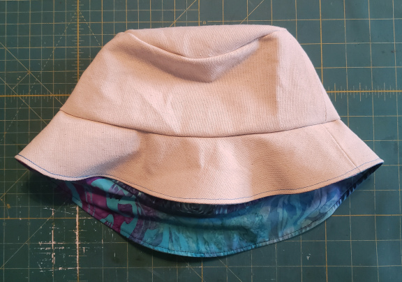 A bucket hat with a cream colored canvas exterior and a blue and purple interior lays on its side.