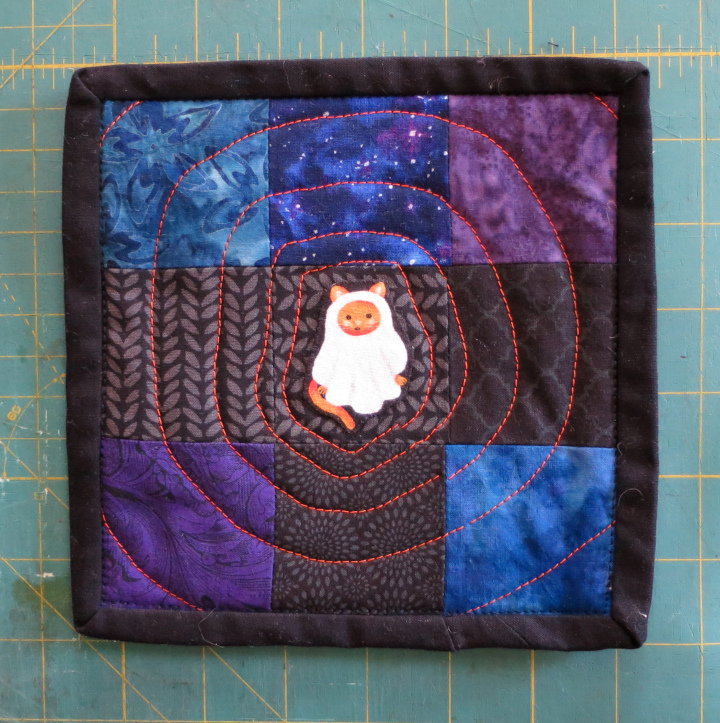 A nine patch quilt block in blue, purple and black with a cat and a ghost costume in the center and orange stitching. The title of this quilt is The Center of Attention.